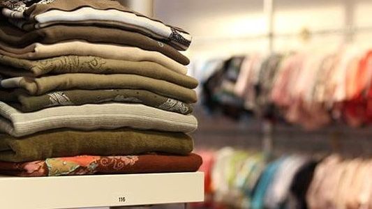 gestapelte Pullober im Vordergrund und hängende Kleidung im Hintergrund.